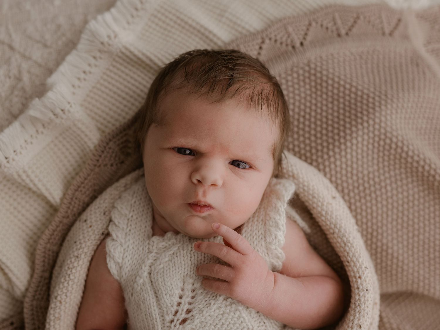 A newborn baby wearing a white knit outfit rests on soft beige blankets while looking at the camera.