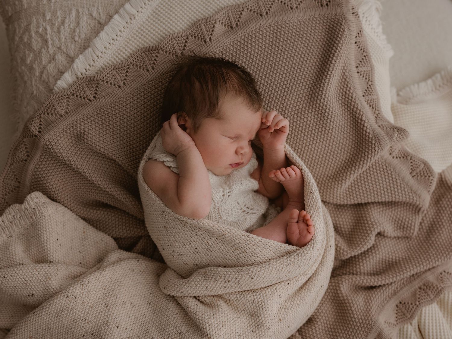 A sleeping newborn baby wrapped in soft beige knitted blankets on textured bedding.