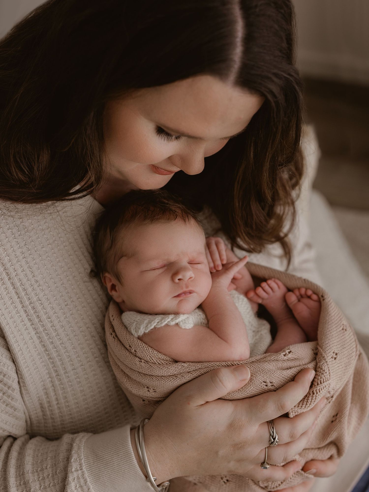 A tender moment shows a parent in a white sweater cradling a sleeping newborn wrapped in a soft beige blanket.