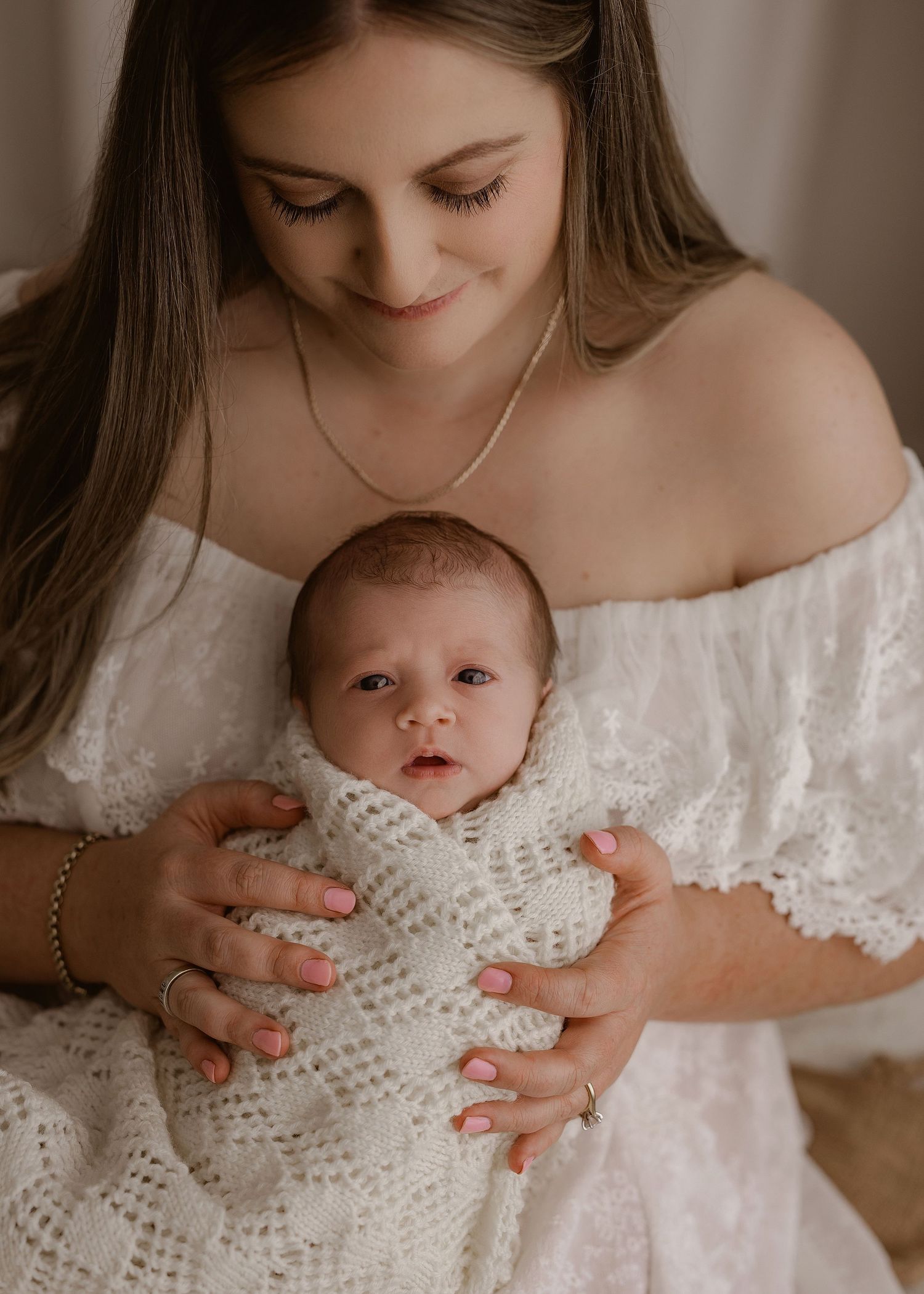 A tender moment shows someone in a white lace dress holding a newborn baby wrapped in a knitted white blanket.