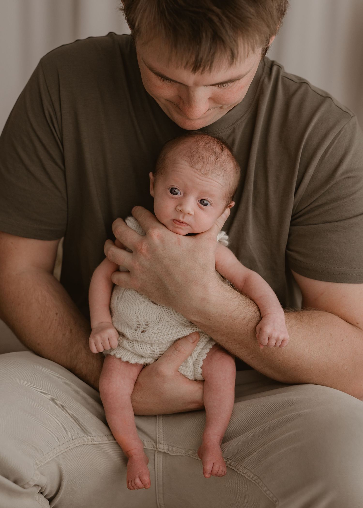 An adult in a brown t-shirt gently holds a newborn baby wearing white knit clothing while sitting on a beige chair.