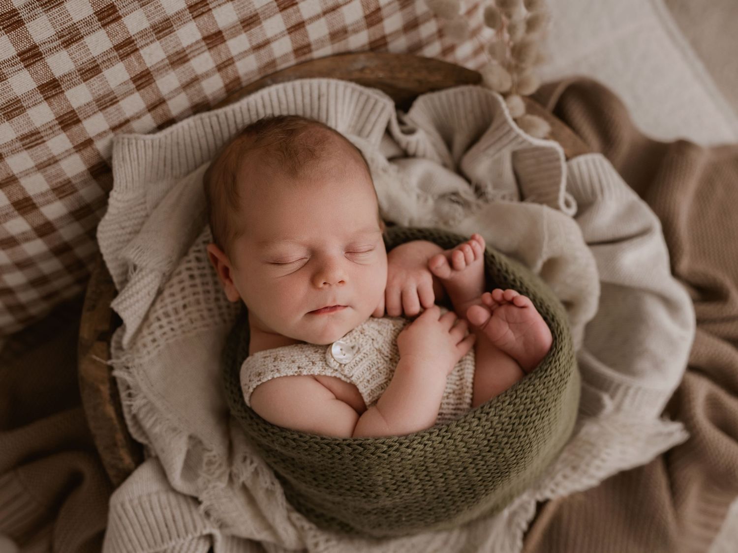 Sleeping newborn nestled in olive green bowl surrounded by soft beige knit blankets in warm neutral tones.