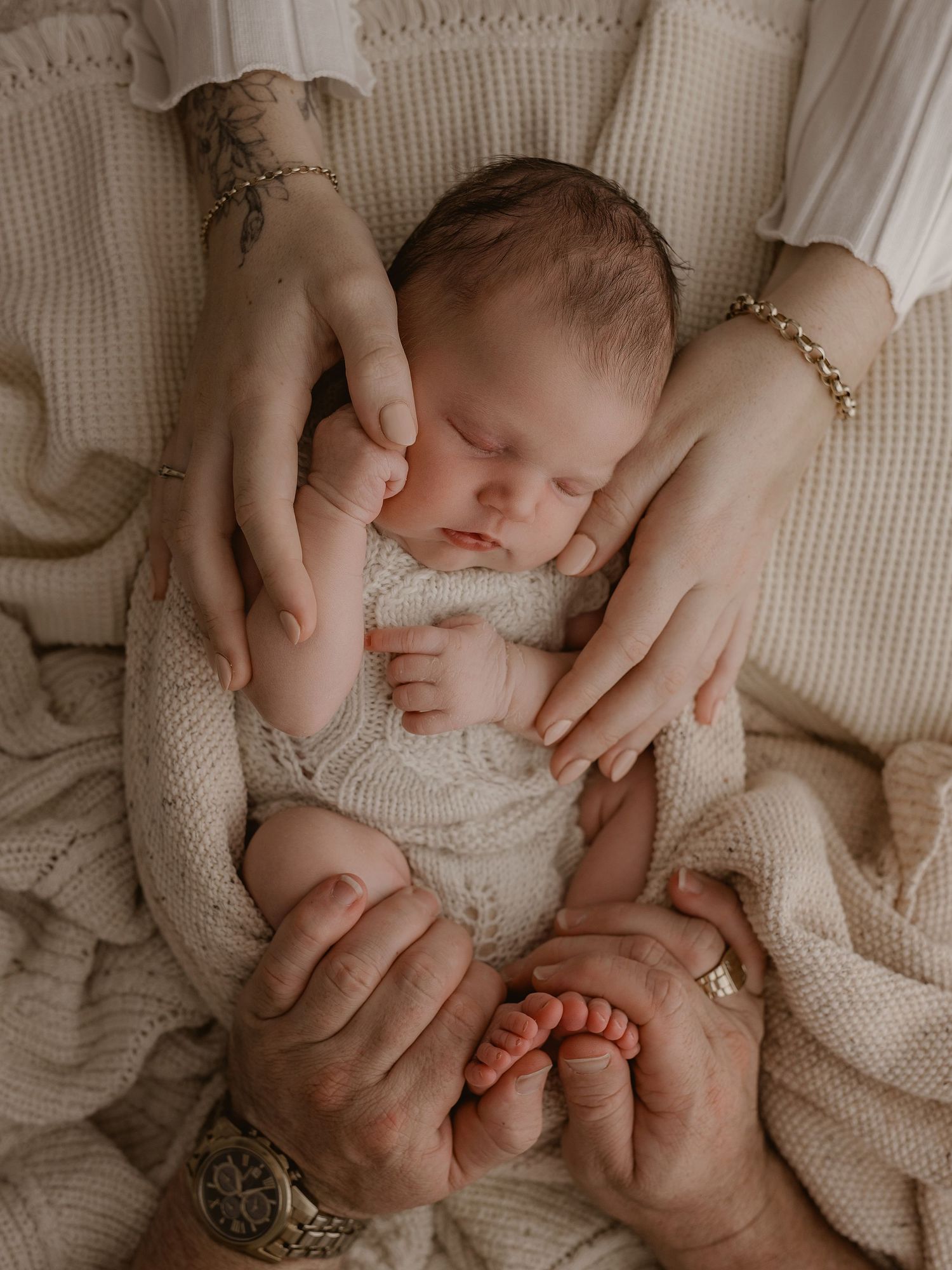 A sleeping newborn baby wearing a white knit outfit is cradled by gentle hands with jewelry and a tattoo visible.