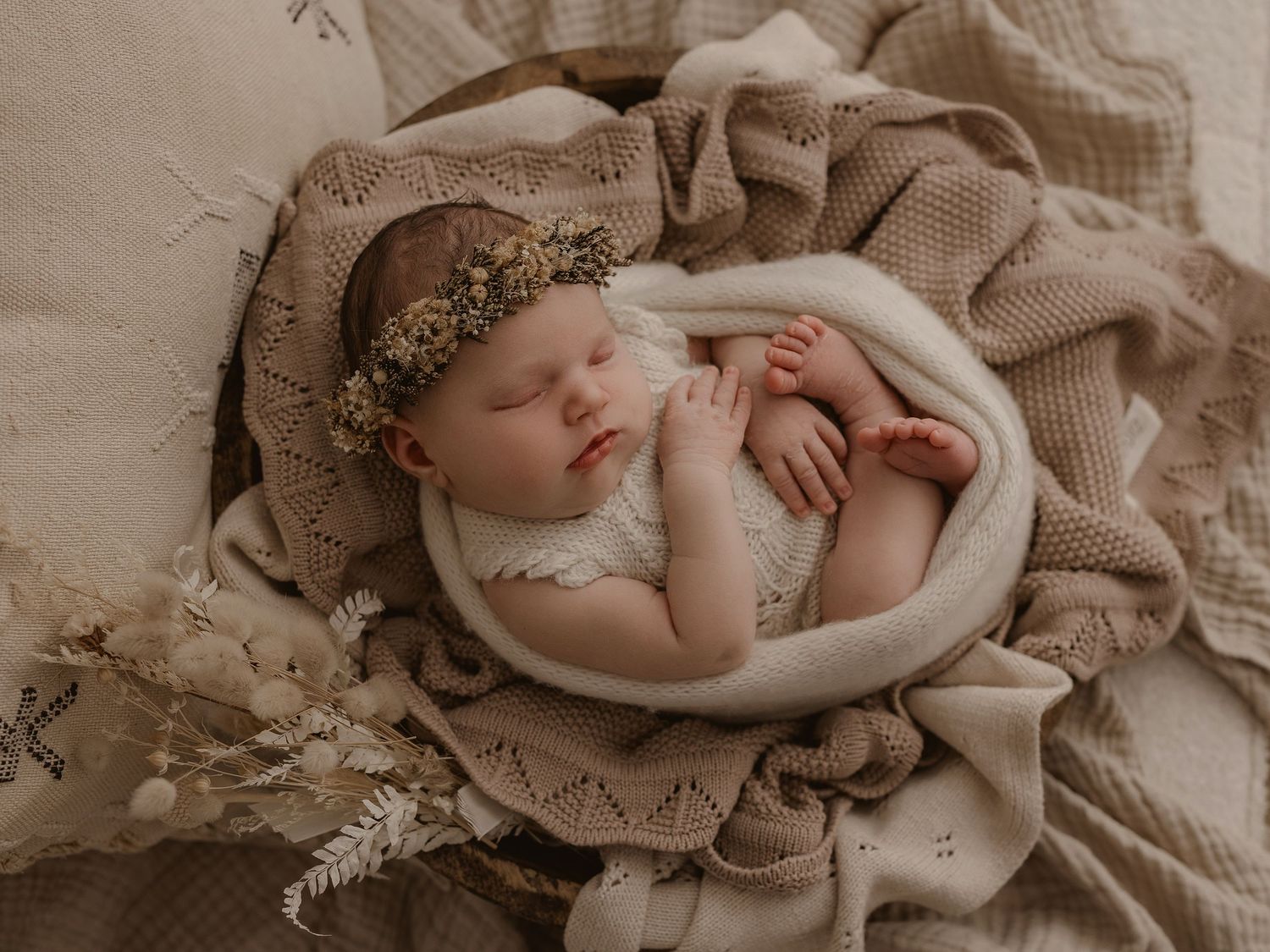 Peaceful newborn baby sleeping in cream knit wrap with floral headband for professional portrait photography.