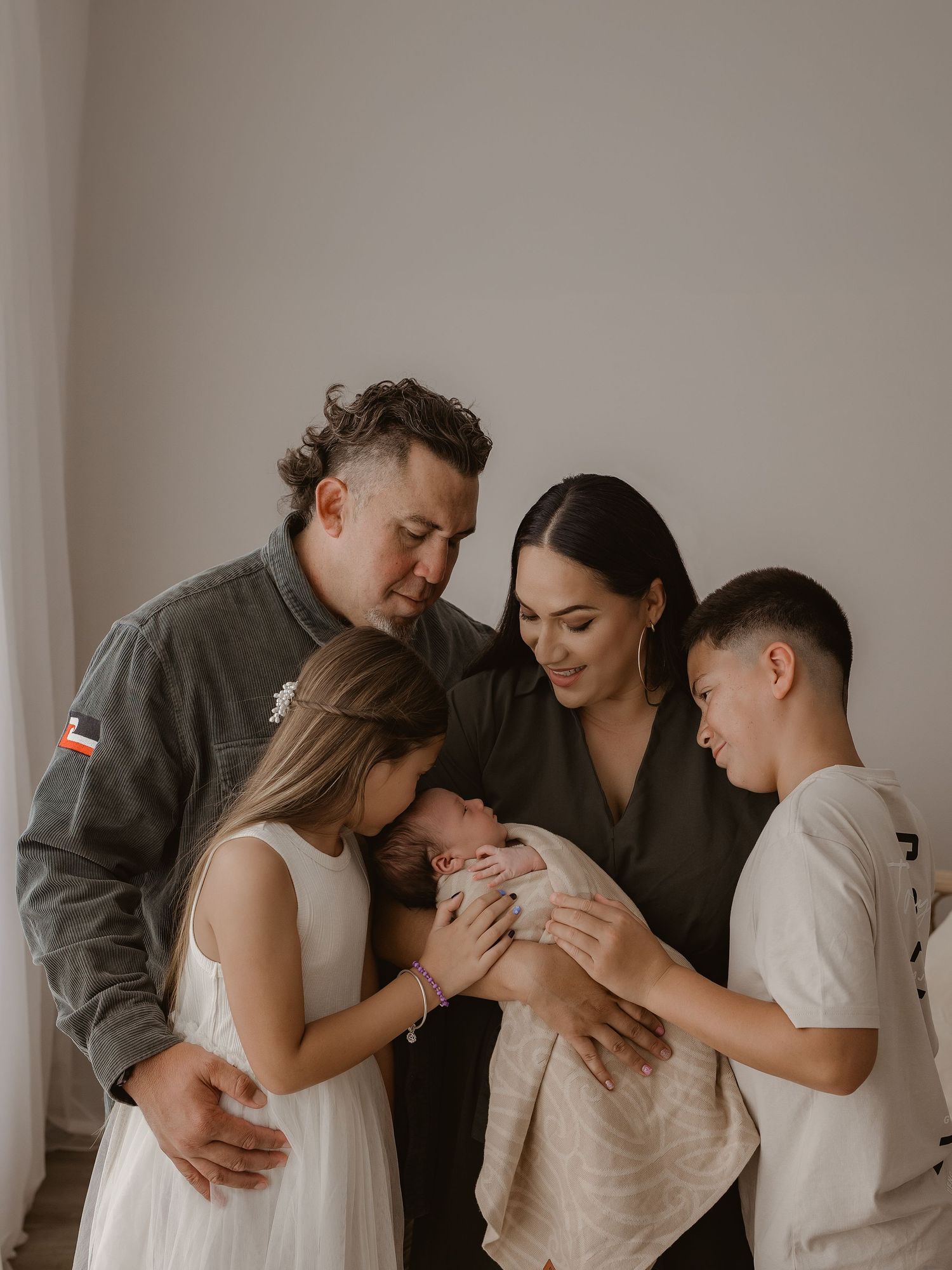 Family gathers together in tender moment with newborn baby in minimalist neutral-toned setting.