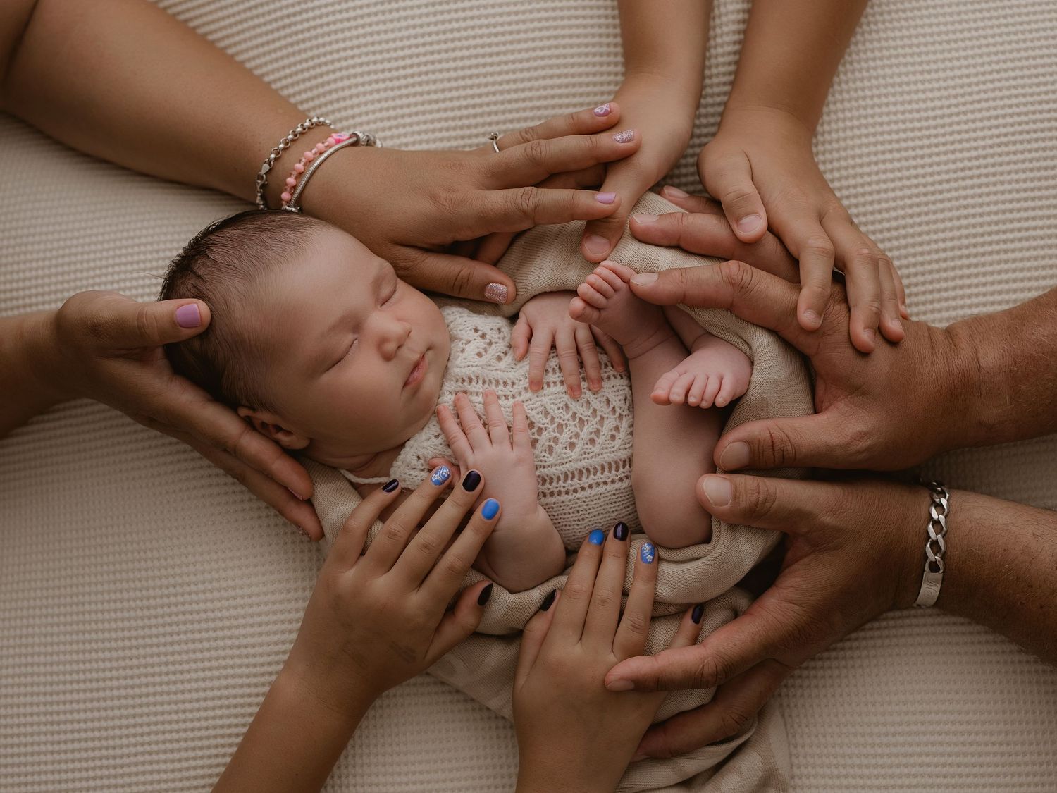 Several hands gently cradle a sleeping newborn baby wrapped in a soft patterned blanket.
