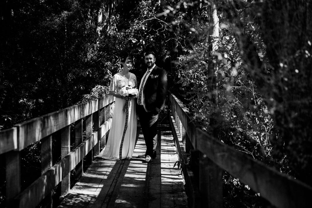 A black and white photo of a couple standing on a rustic wooden bridge in the woods.
