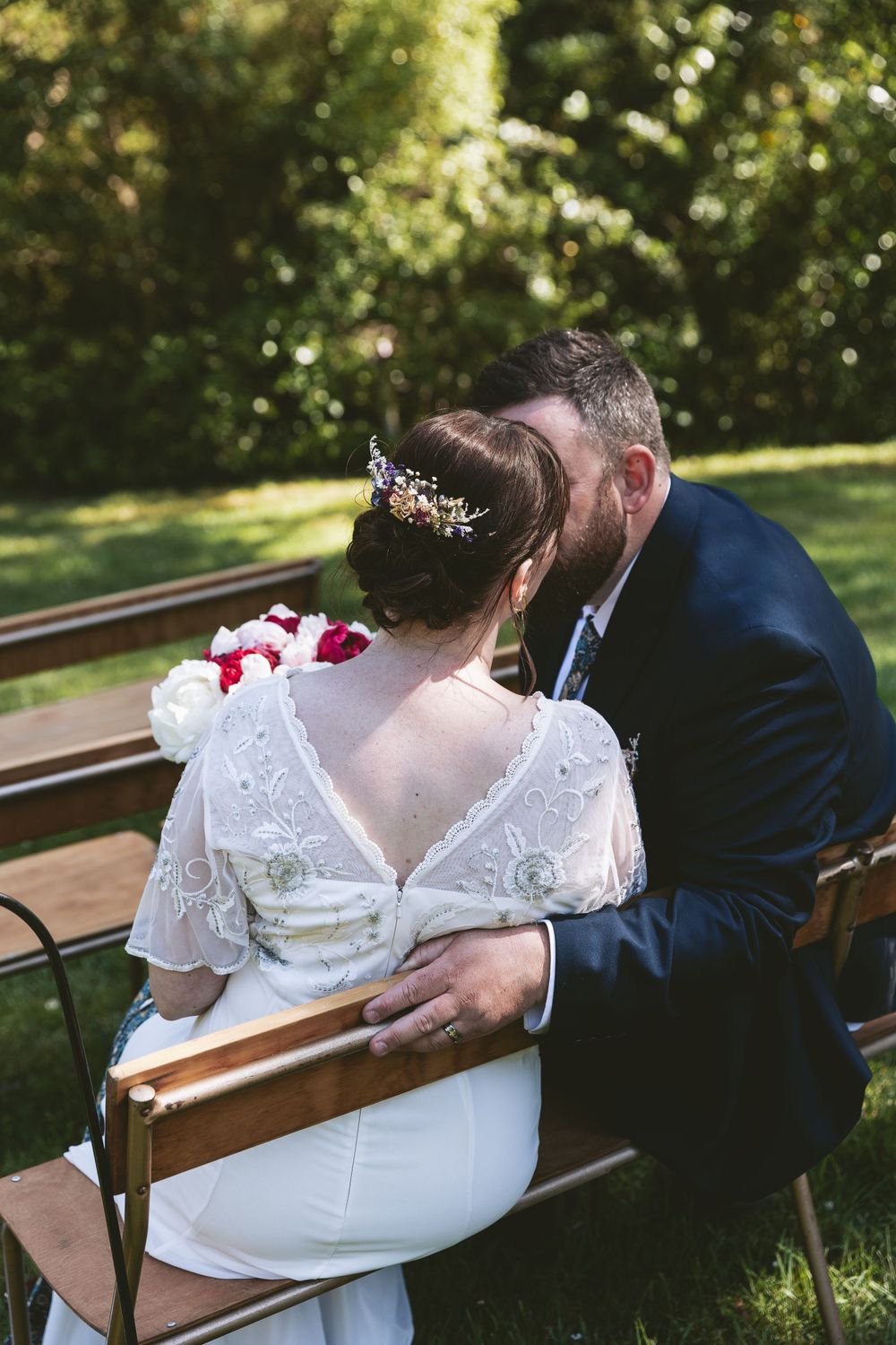 A tender wedding moment photographed on a wooden bench in a garden setting.