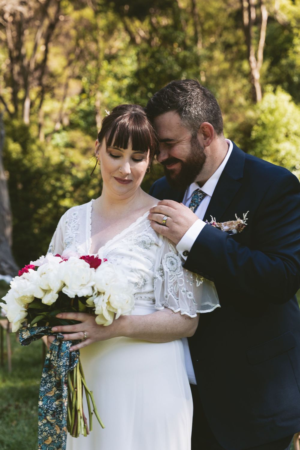 A wedding couple shares an intimate moment outdoors while holding a white and red floral bouquet with blue ribbon.