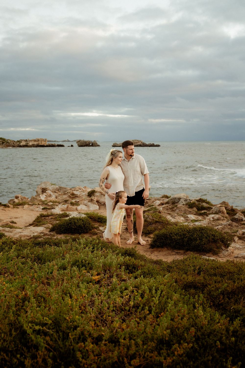 A couple stands together on a rocky coastal cliff overlooking the ocean during golden hour at sunset.