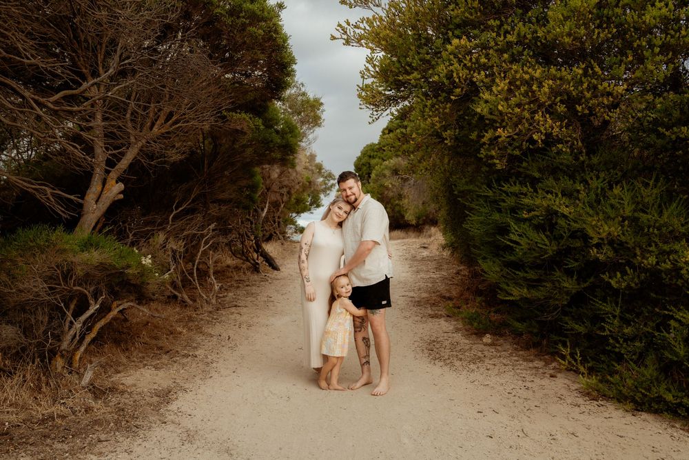 A couple embraces on a sandy nature trail surrounded by green foliage on a sunny afternoon.