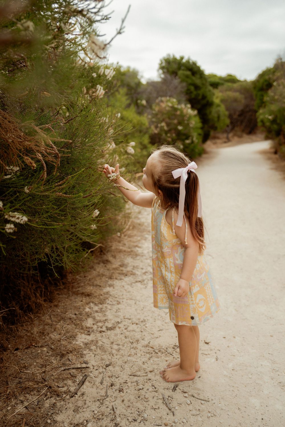 Young figure in floral dress reaching out to touch flowering bushes along a dirt path on an overcast day.