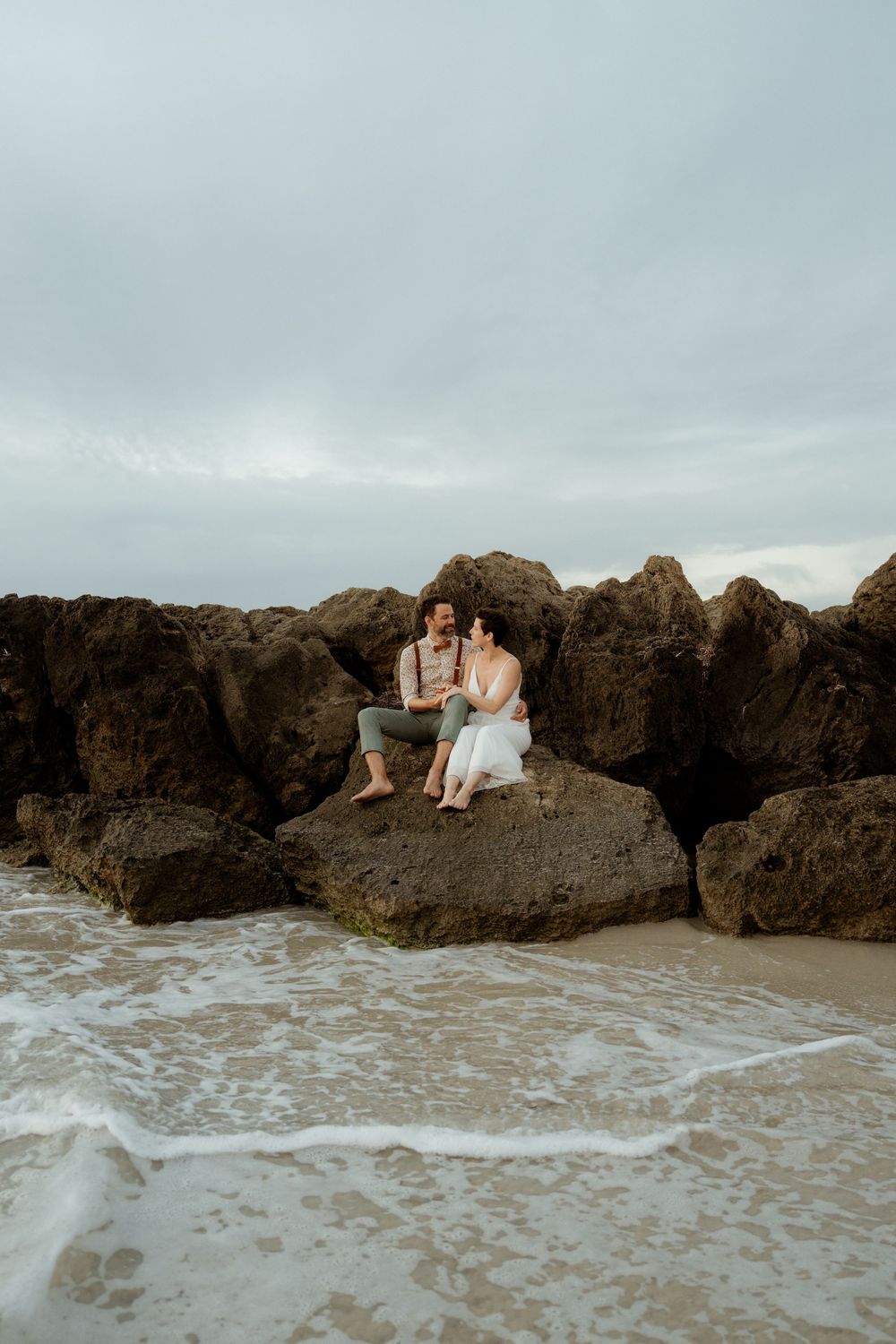A couple sits together on coastal rocks as ocean waves crash against the shore during an overcast evening.