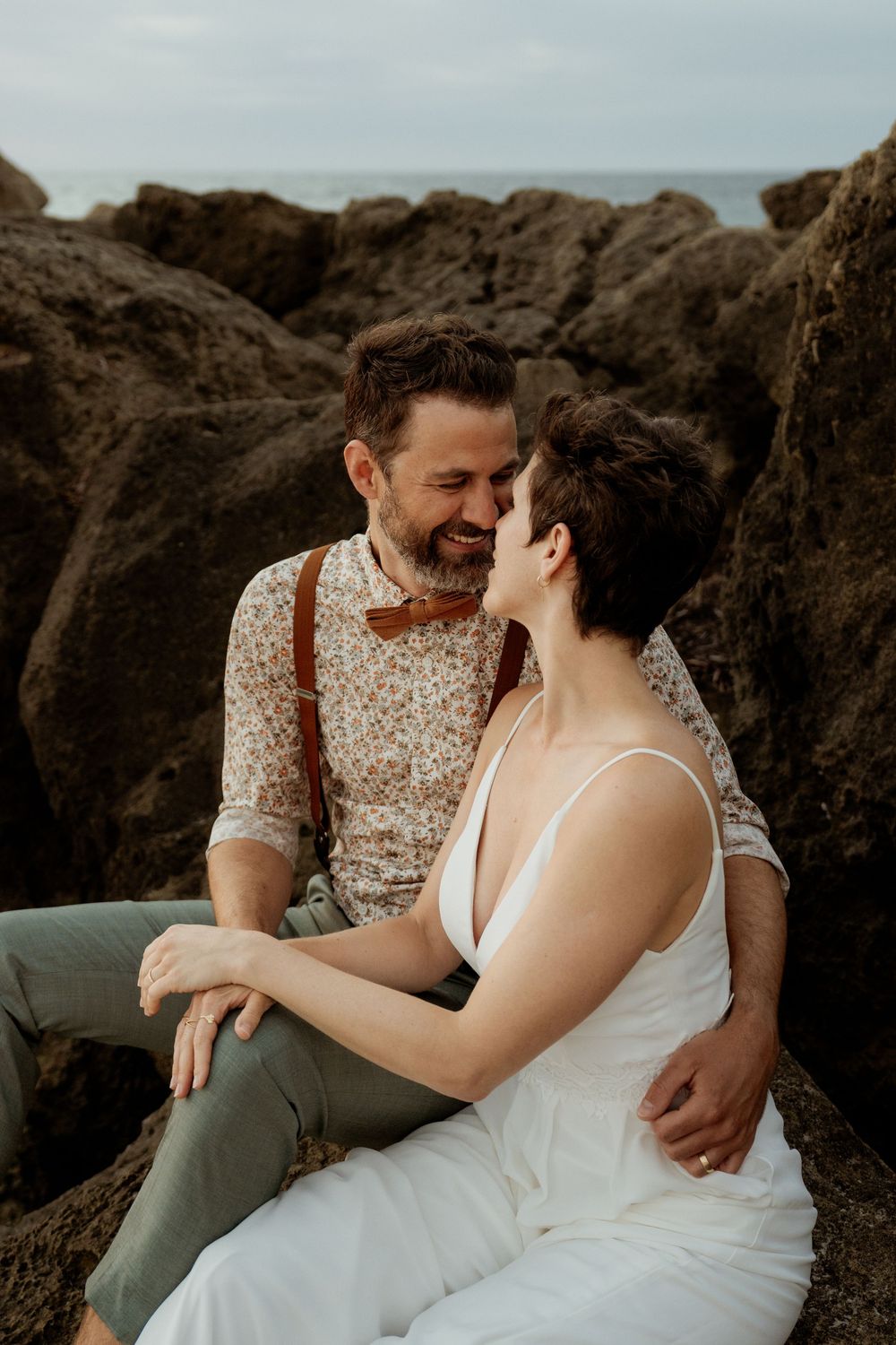 A couple shares an intimate moment while sitting on coastal rocks at sunset in wedding attire.