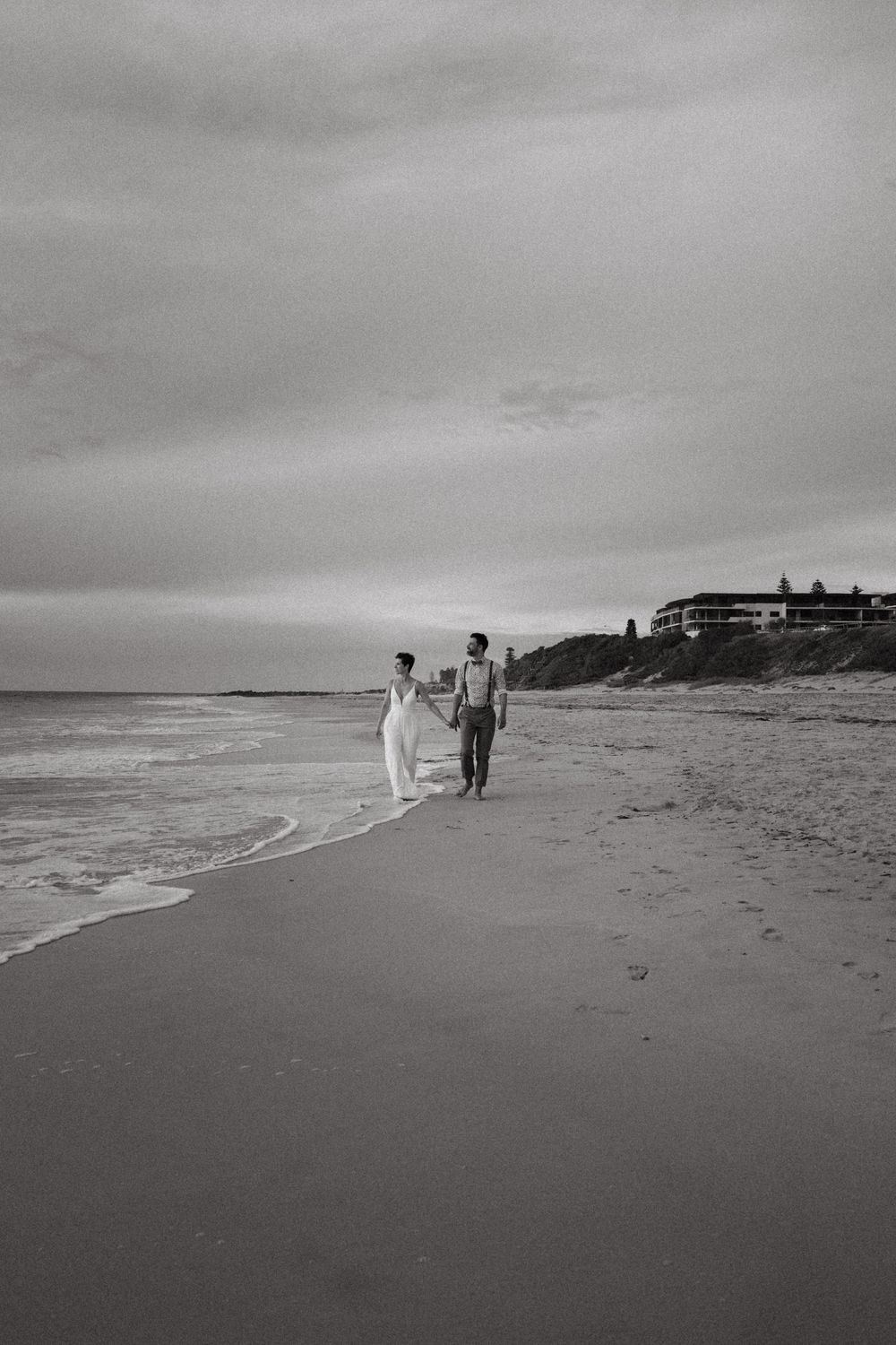 A couple walks along a serene beach at sunset with buildings visible on the hilltop in black and white photography.