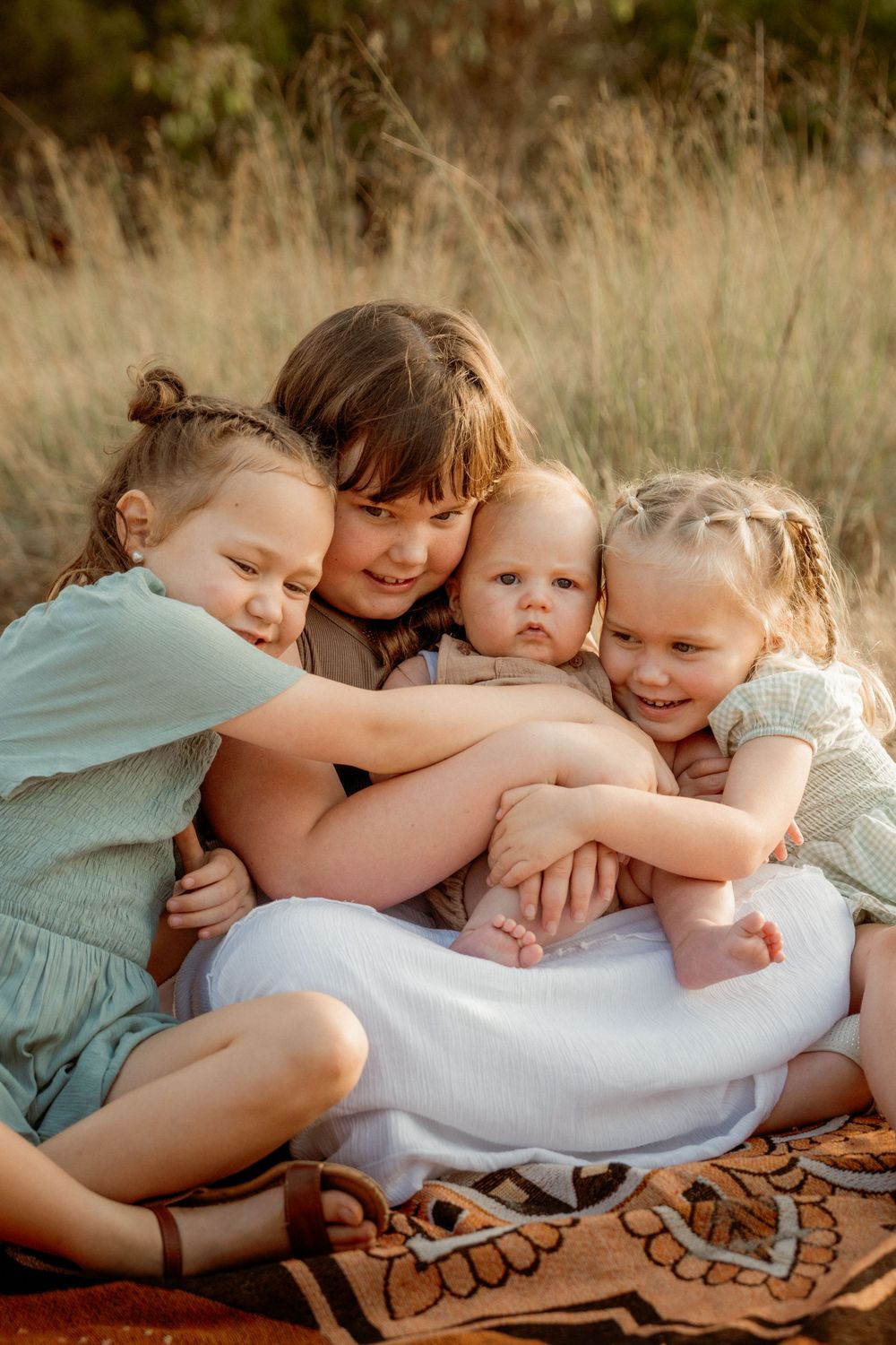 Four children cuddle close together on a patterned blanket in a field of tall grass during golden hour.
