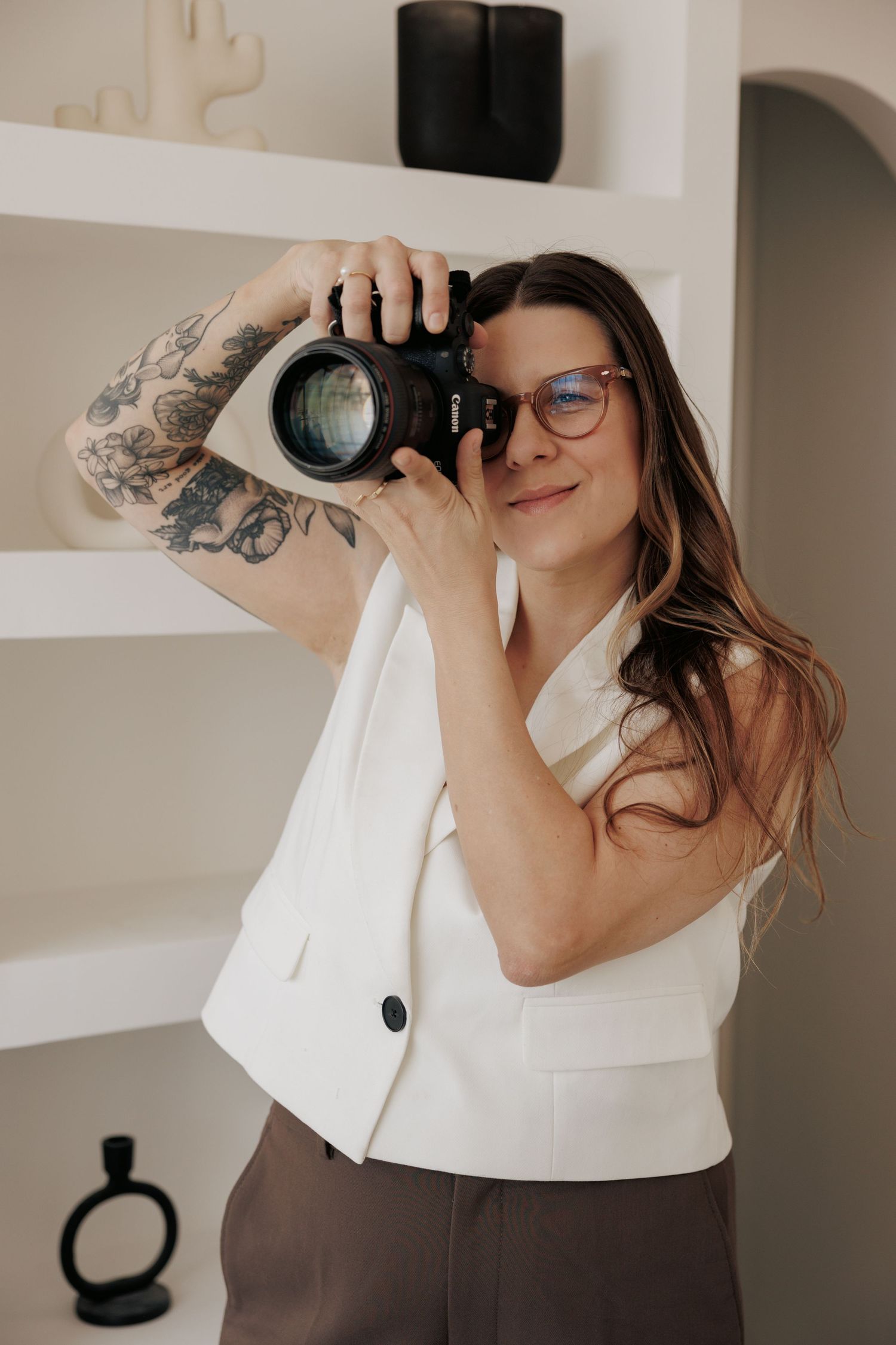 Professional photographer in a white vest top showing off artistic tattoos while holding a camera.