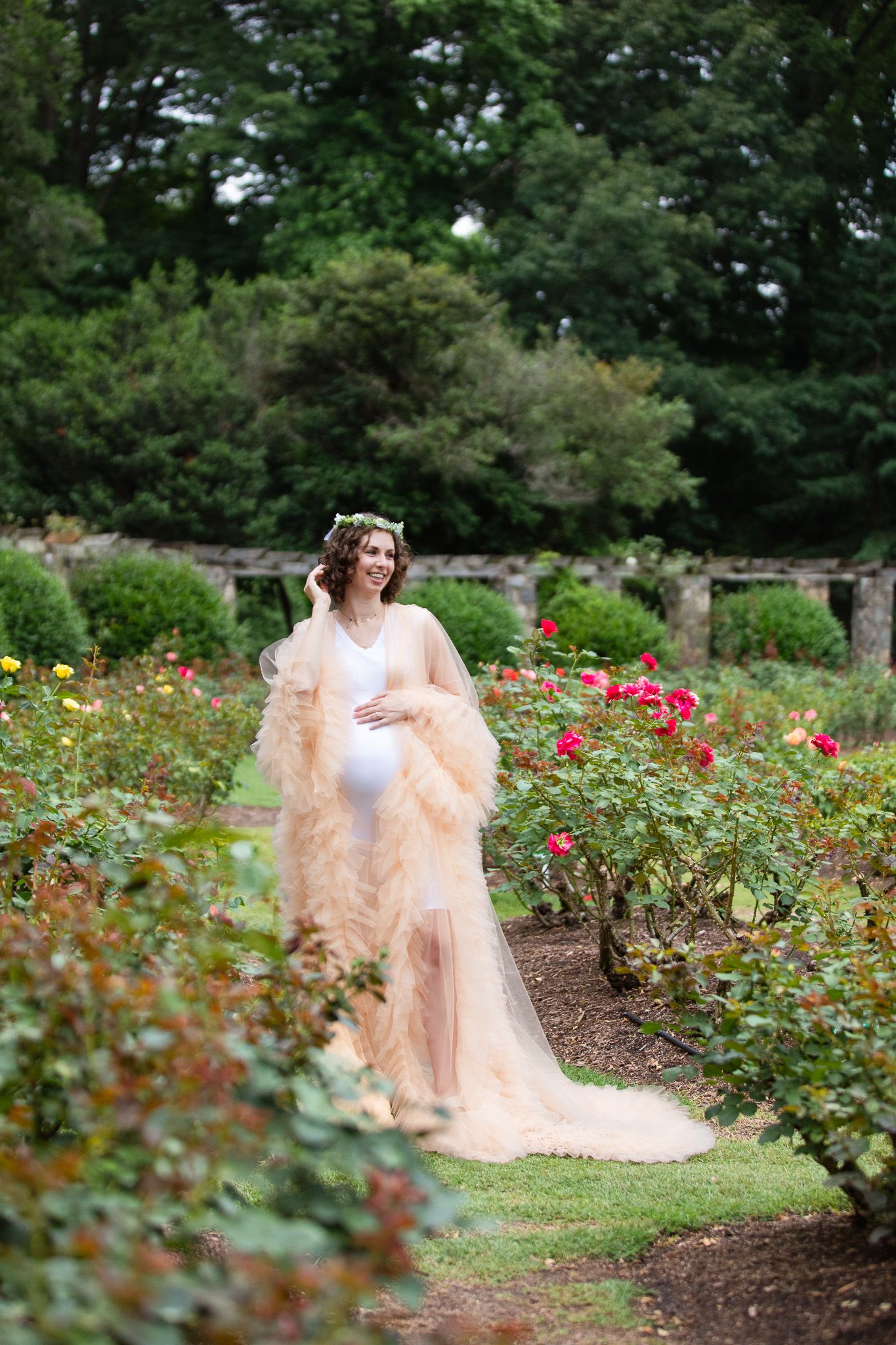 A romantic garden maternity photo features a flowing peach gown among blooming roses and greenery.
