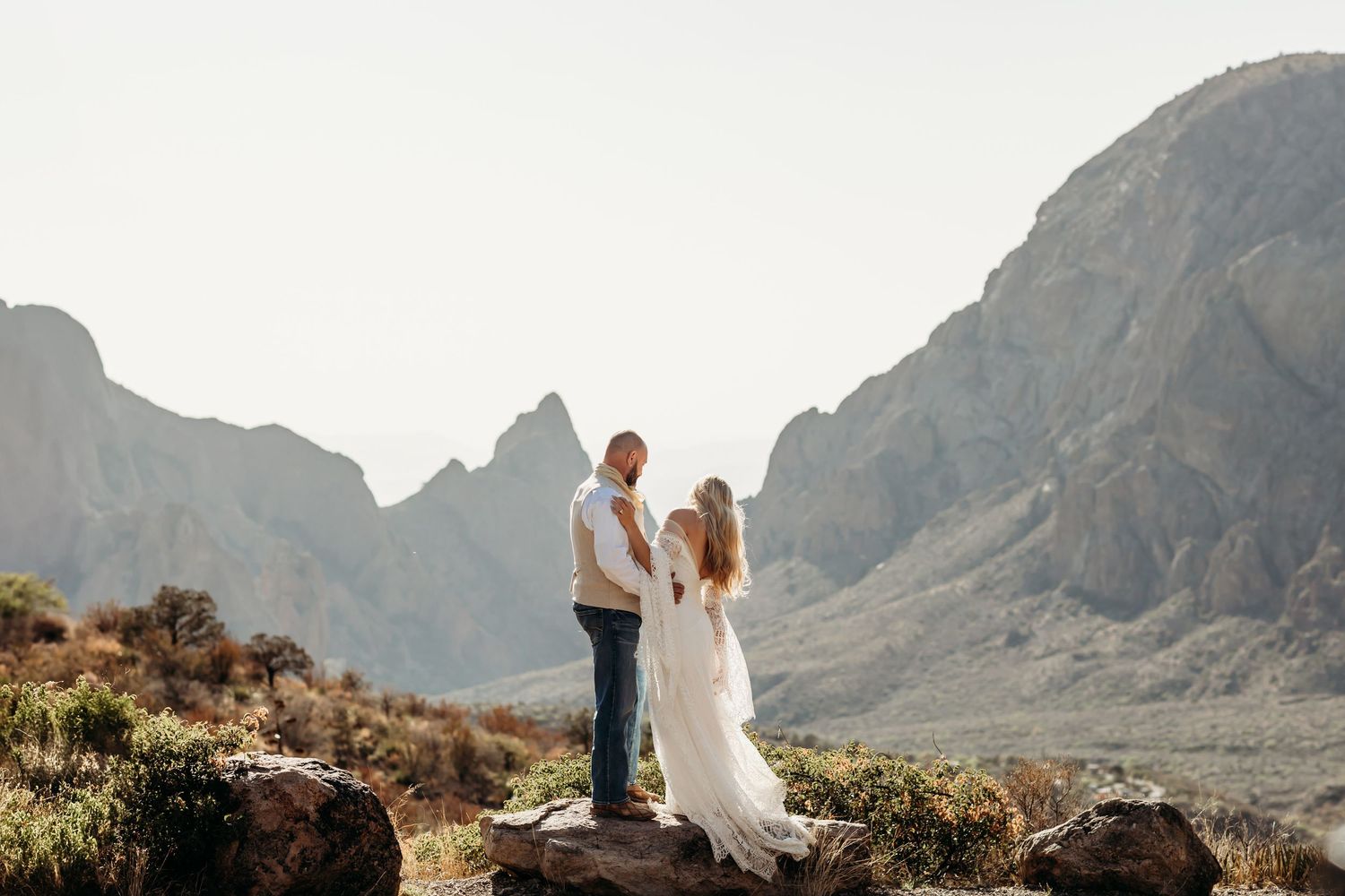 Couple in wedding attire stands on rocky overlook with dramatic mountain peaks in background during sunset.