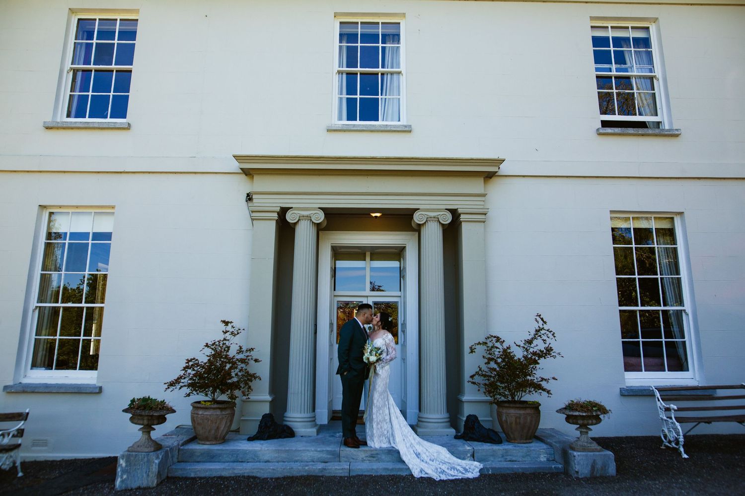 A couple stands between white columns on their wedding day, with the bride holding a white and green bouquet.