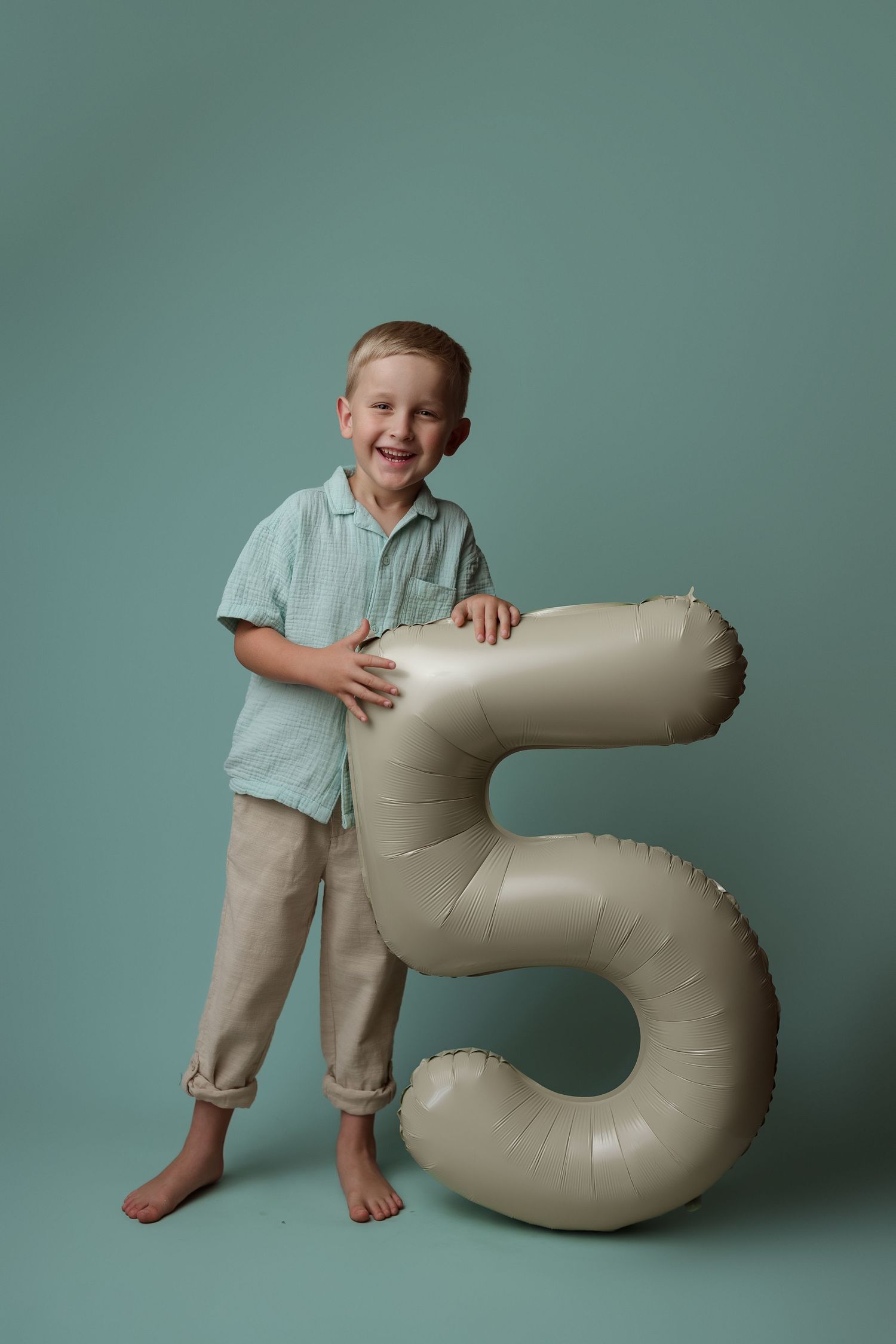 A child in a mint green polo shirt stands barefoot next to a large inflatable number 5 against a teal backdrop.