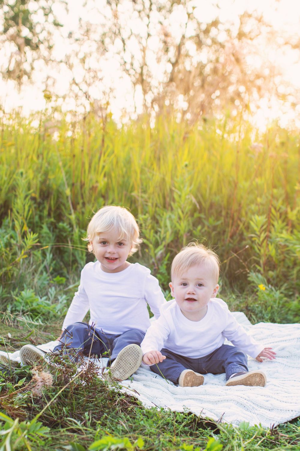 Two babies in matching outfits sit on a white blanket in a sunlit meadow.