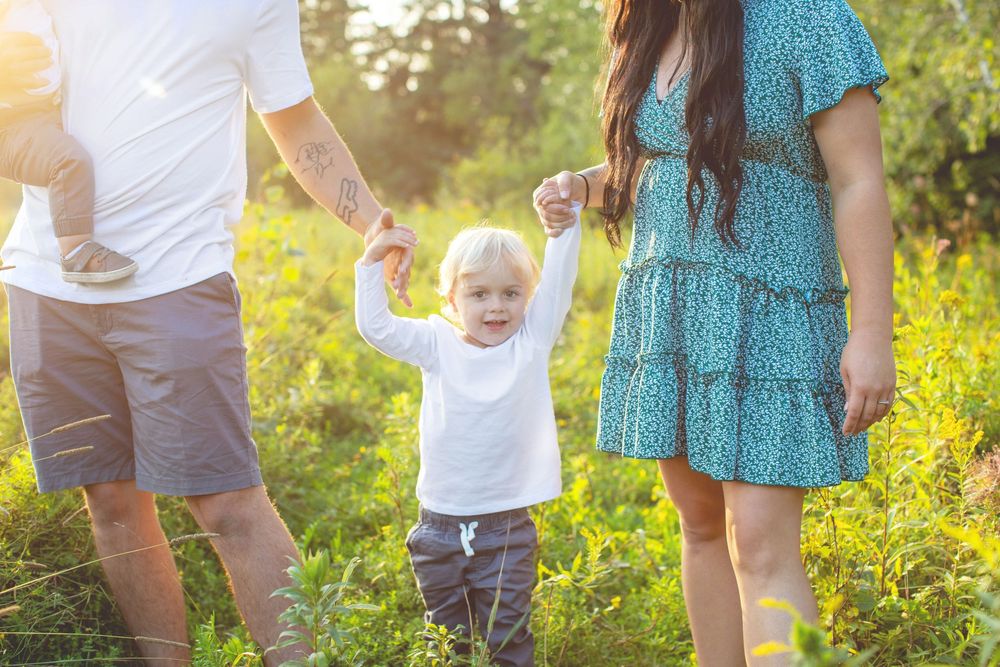 A child in a white shirt walks through a sunlit wildflower meadow holding hands with adults on a summer evening.