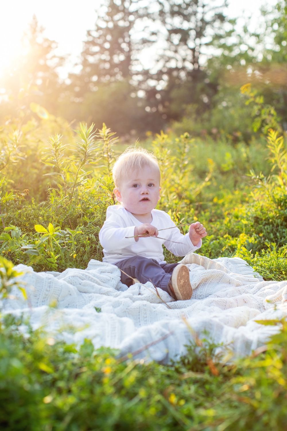 Baby in white outfit lying on blanket outdoors during golden sunset with green foliage in background.