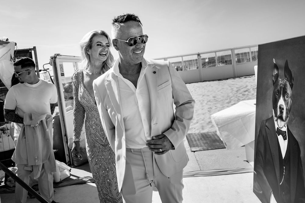 Stylish couple in light summer attire walking on a beach with framed artwork visible in background.
