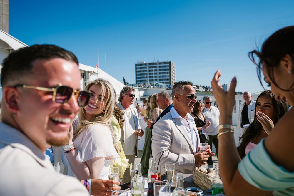 People socialize and enjoy drinks while standing outdoors on a sunny day against a bright blue sky.