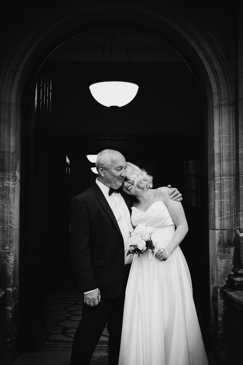 A black and white wedding photo of a couple laughing together under an arched doorway with an overhead light.