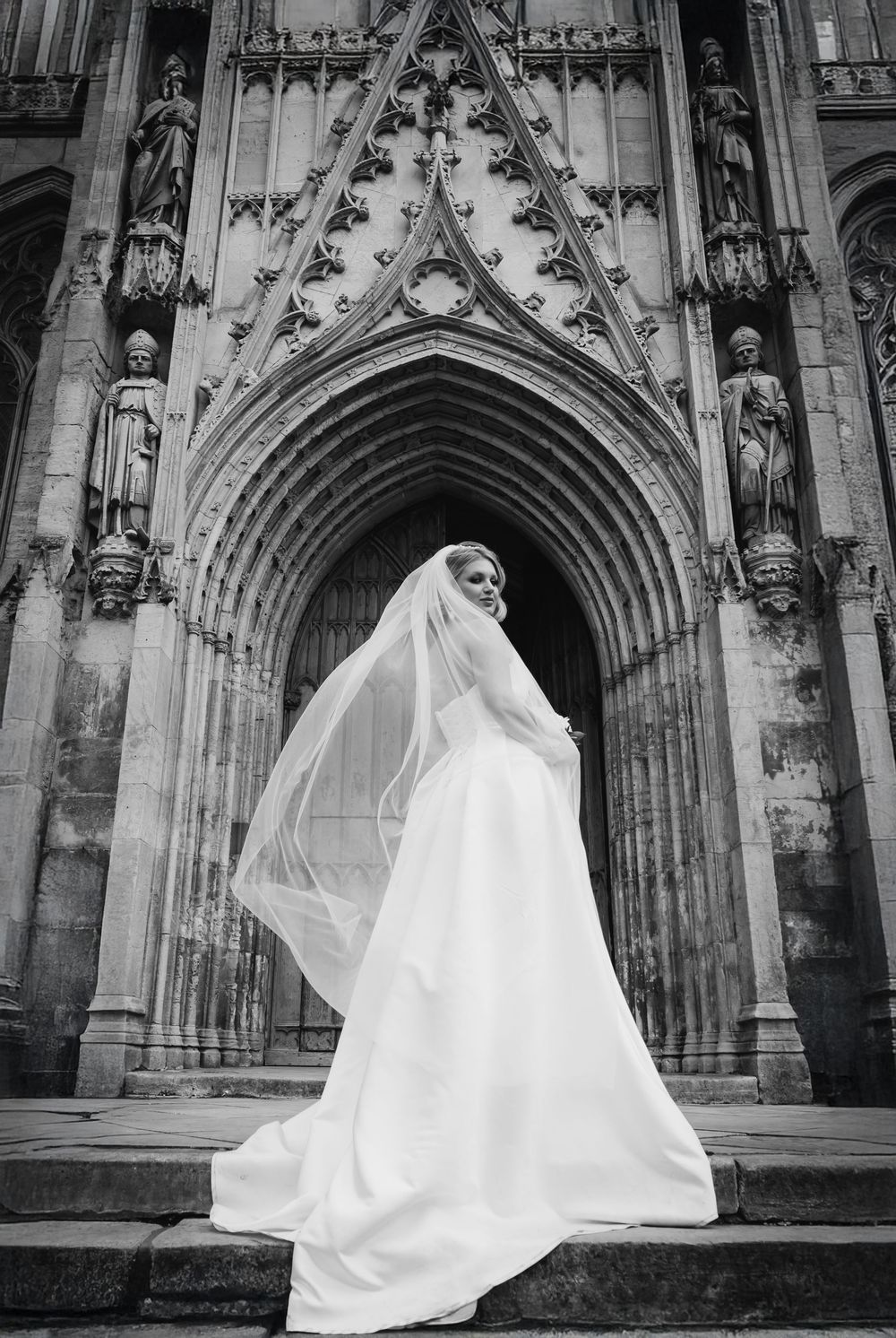 A bride in a flowing white wedding dress and veil stands at a grand Gothic cathedral entrance with ornate stone carvings.