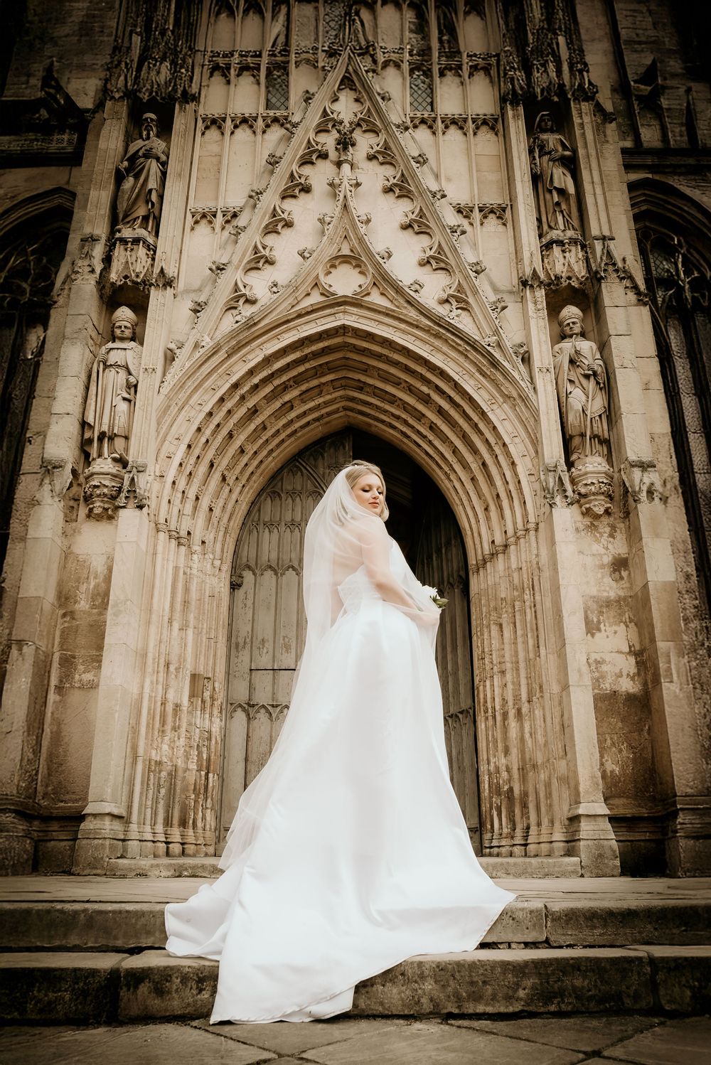 Bride in flowing white dress poses dramatically against ornate Gothic church archways in sepia tones.
