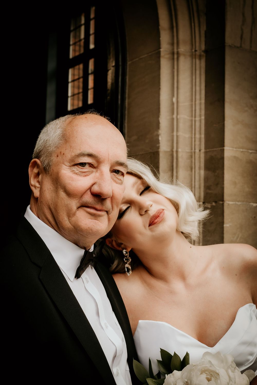 A romantic bridal couple shares intimate moments while posing for wedding portraits against a stone building backdrop.