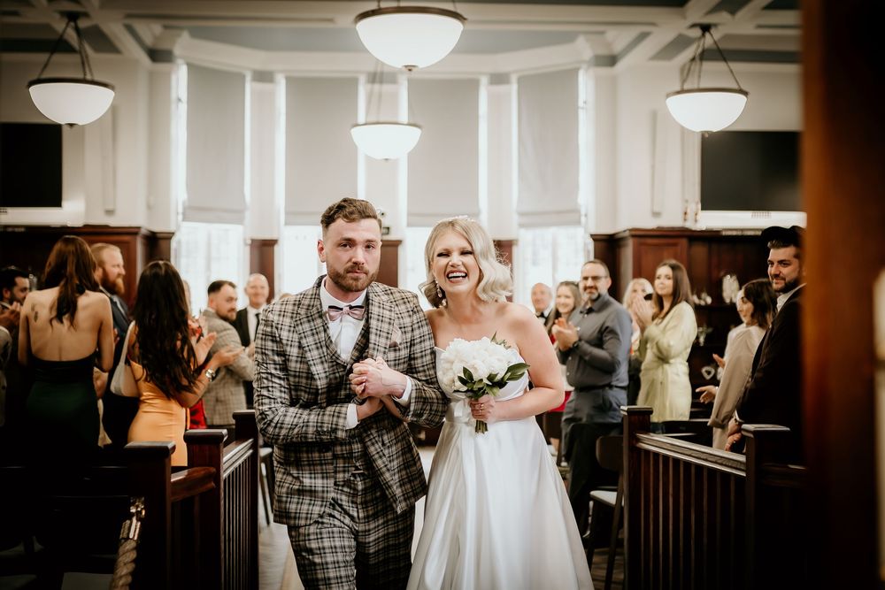 A couple walks down the aisle during their wedding ceremony as guests celebrate in an indoor venue with pendant lighting.