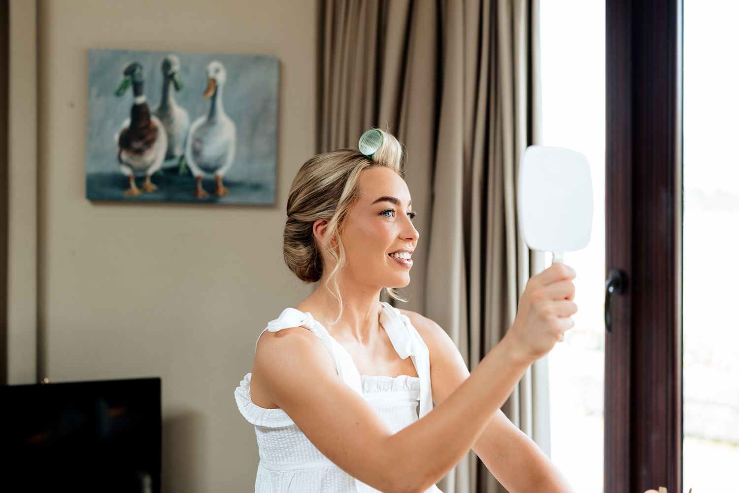 A person in a white garment holds a glass of champagne while preparing for an event by a window.