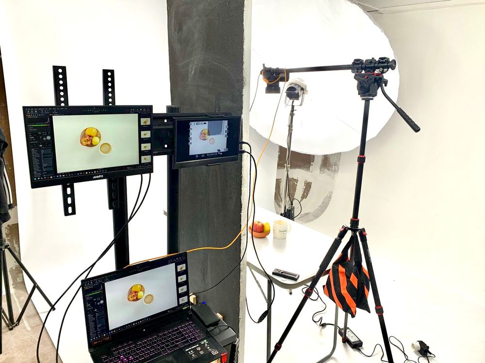 Photography studio setup with computer monitors and lighting equipment mounted on stands against a white wall.