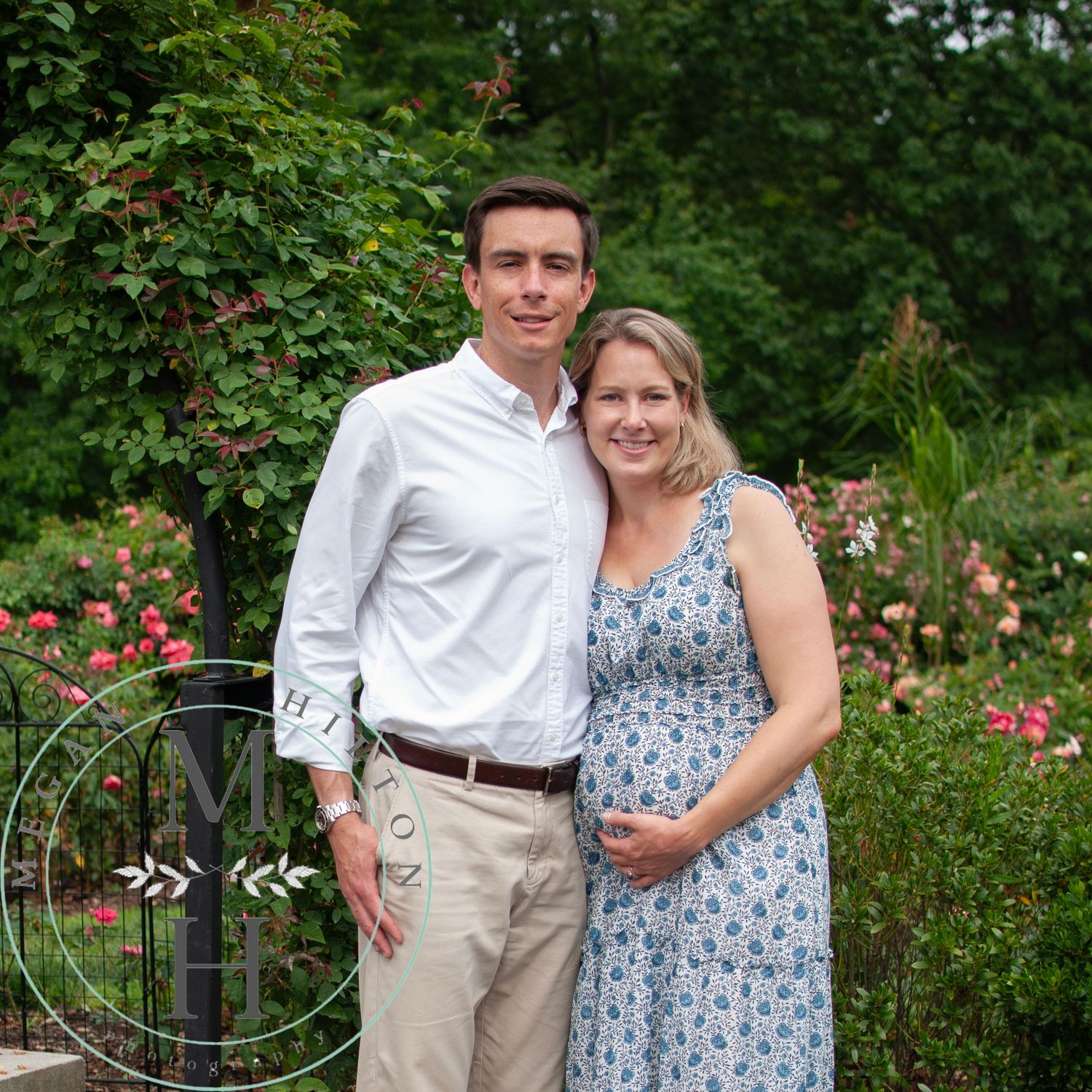 Couple poses in garden setting with blooming pink roses and greenery, woman wearing floral dress.