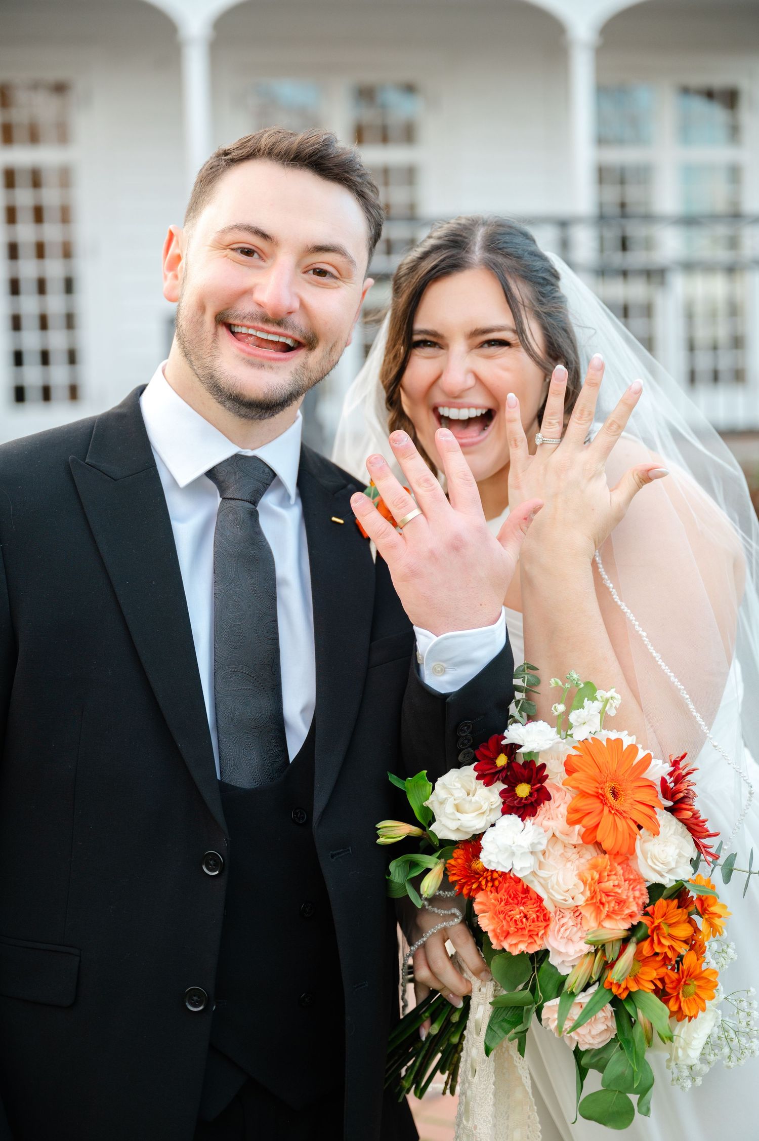 Happy newlyweds show off their wedding rings while holding a vibrant orange and red bouquet outside a columned building by NYC photographer
