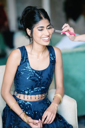 A person in a blue sequined outfit smiles while having makeup applied during wedding preparations.
