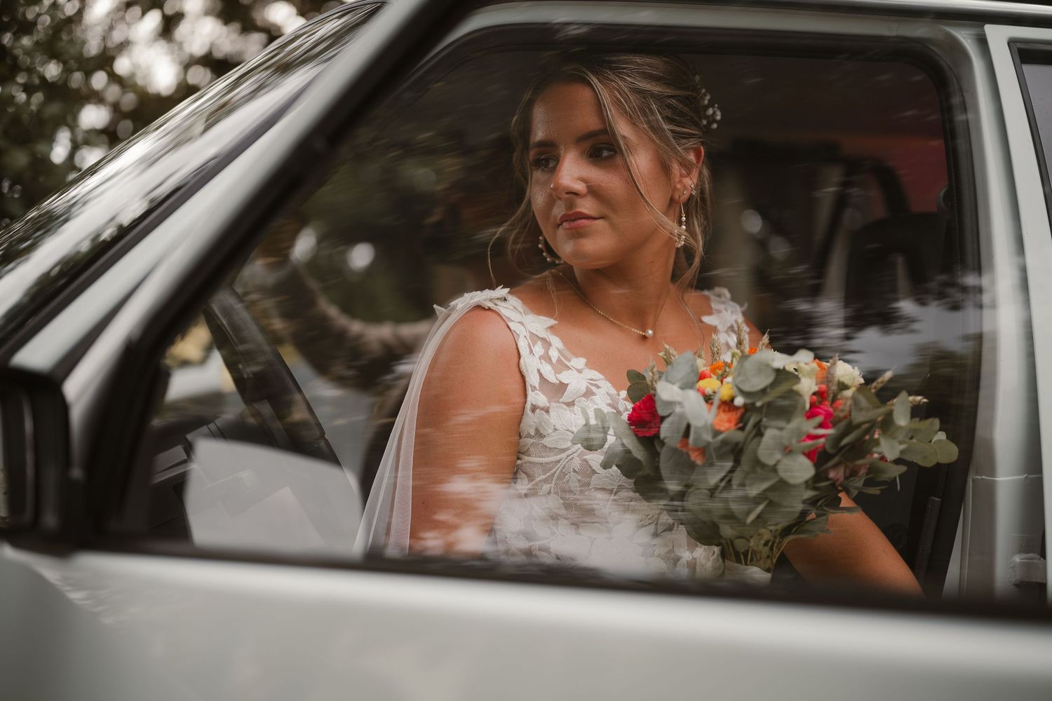 Beautiful bride in white lace dress sits in car with a floral bouquet of pink and whites on her wedding day.