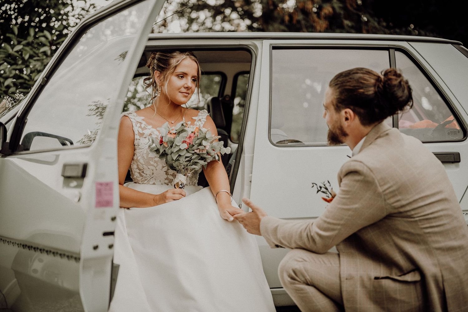 Vintage inspired wedding scene with couple and classic car outdoors in warm natural lighting.