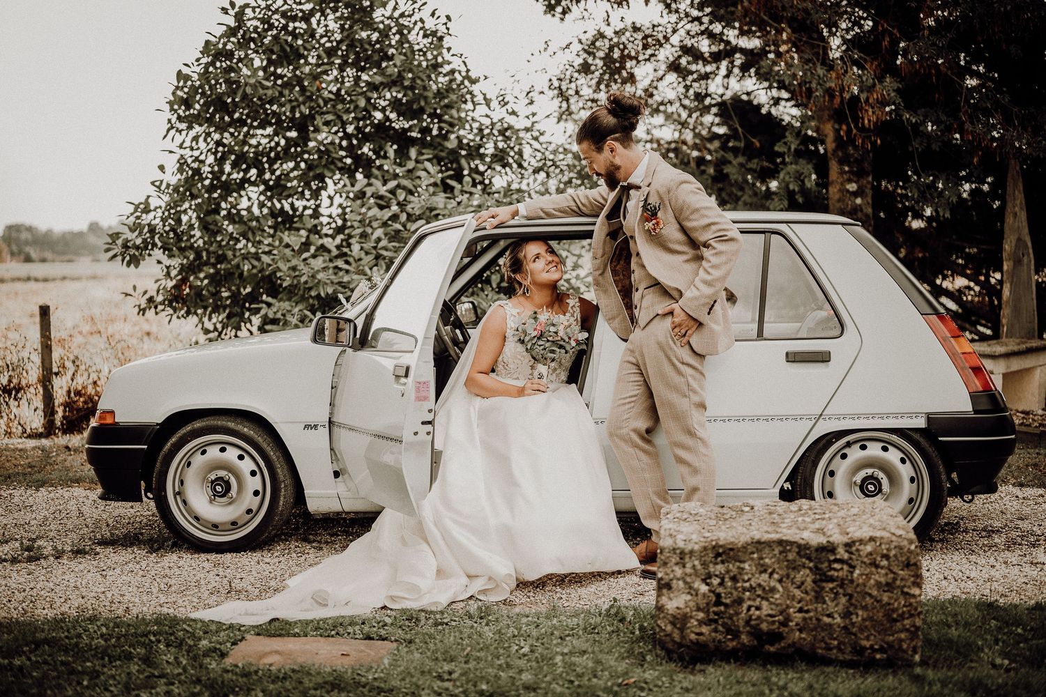 Bride in flowing white dress poses with vintage car in romantic countryside setting.