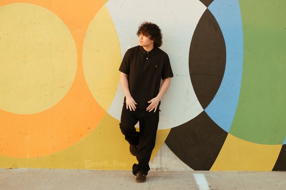 Someone in black attire leans against a colorful geometric mural wall featuring circles in orange, yellow and blue tones.