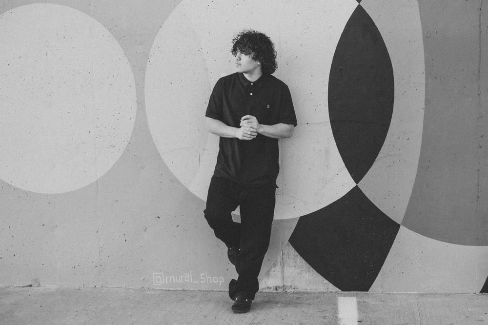 A person in all black attire leans against a black and white geometric wall mural.