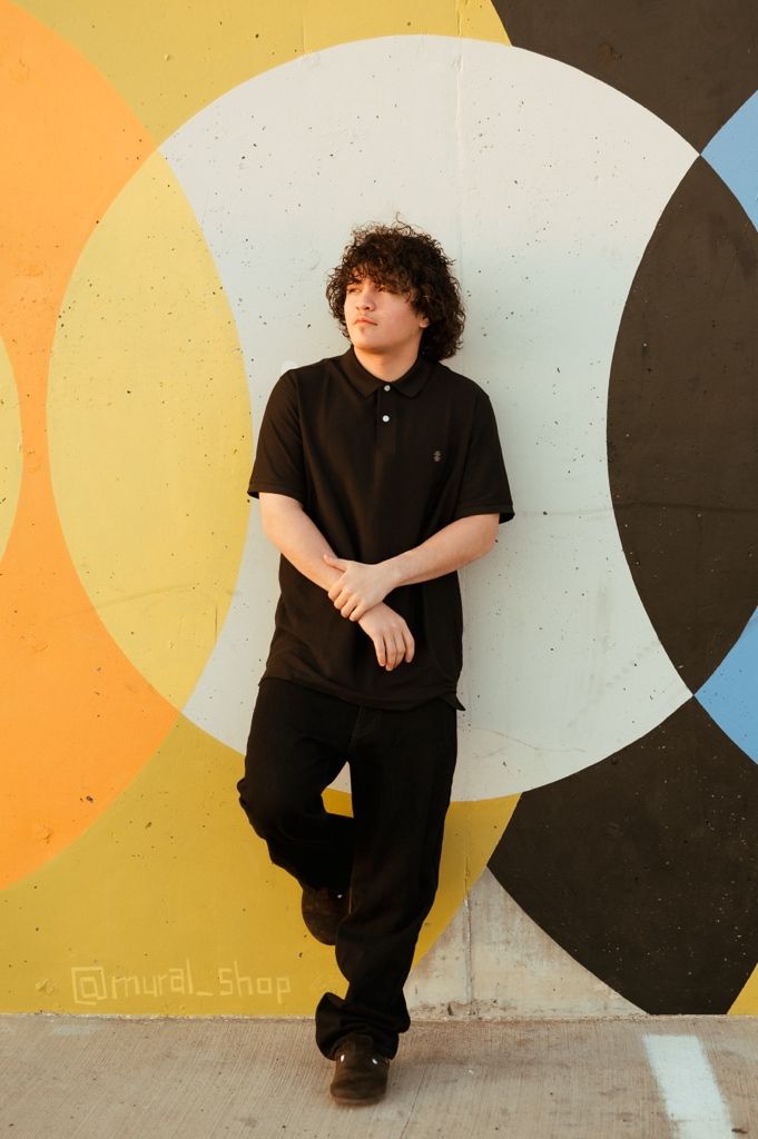 Person in black polo shirt and pants leans against a wall with colorful geometric circles painted as background art.