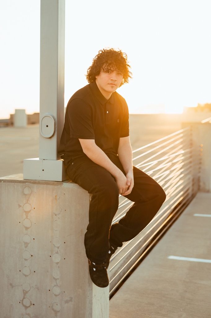 A silhouetted figure in dark clothing sits on a concrete ledge during golden hour sunset on an urban rooftop.