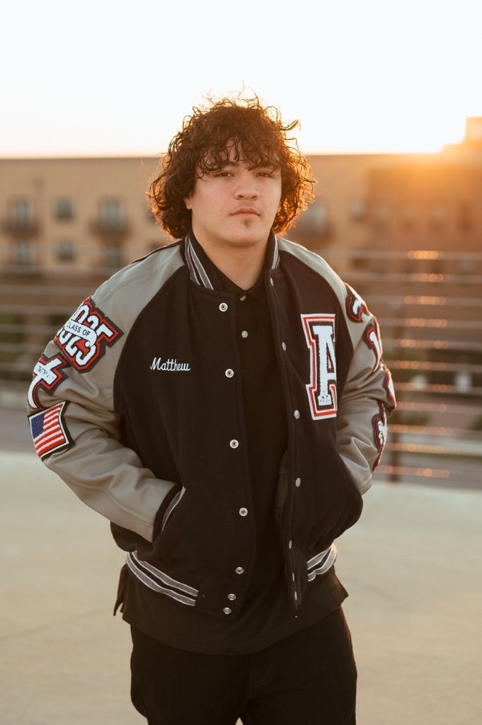 A person wearing a black and gray varsity letterman jacket poses outdoors during golden hour sunset.