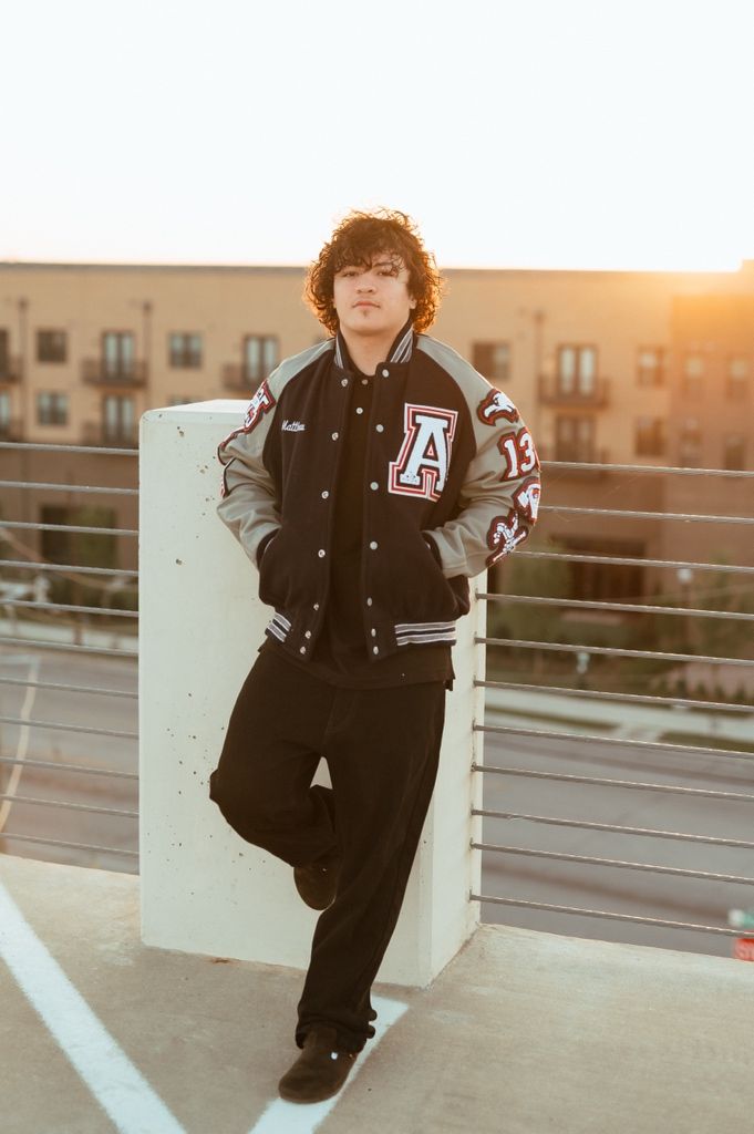 Varsity letterman jacket wearer stands at sunset on rooftop parking deck leaning against concrete barrier.