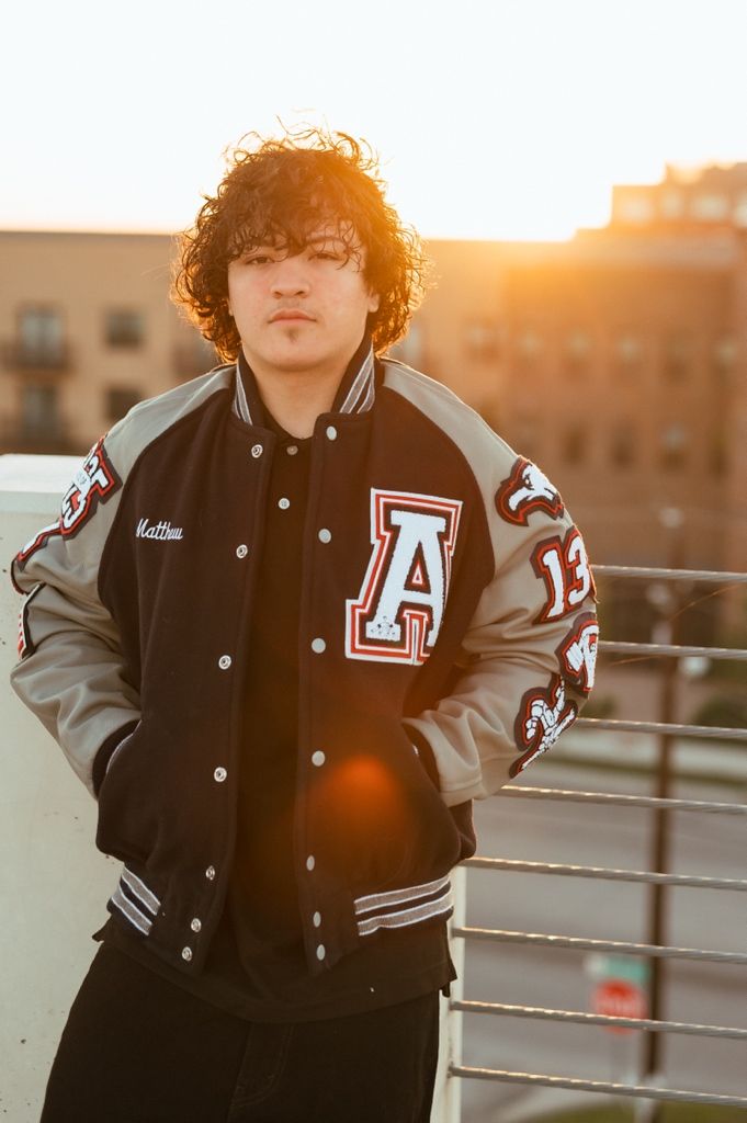 A person in a varsity letterman jacket poses outdoors at sunset with a sun flare behind them.